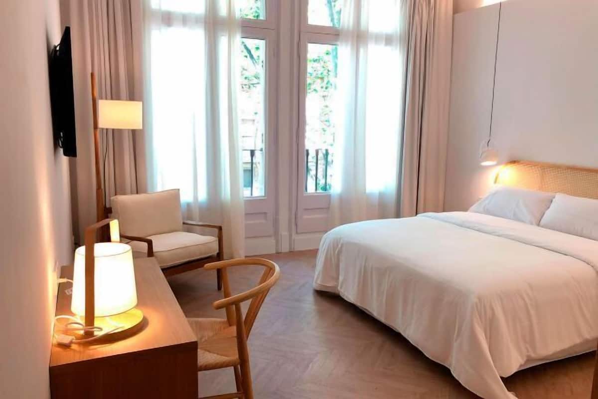 A hotel room with a bed, a desk with a lamp, a chair, and a TV mounted on the wall.