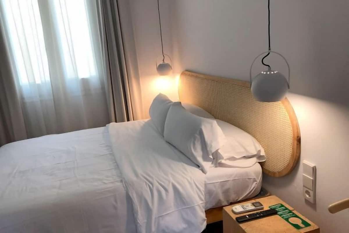 A hotel room with a bed, pillows, a nightstand with a remote control, and a wall socket.
