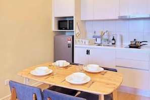 Een compacte keuken met een magnetron, koelkast en een klein spoelbakje. Er staat een houten eettafel met stoelen en de keuken heeft witte kastjes en een glazen aanrechtblad.