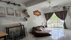Een ruime woonkamer met een bank, een eettafel met krukken, een plafondventilator en een groot raam met gordijnen.
