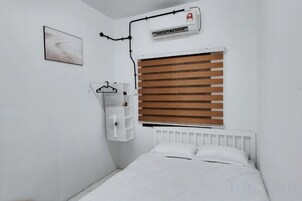 Un dormitorio con una cama blanca, un radiador, una ventana con persianas y una estantería en la pared.