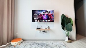 A living room with a flat-screen TV mounted on the wall, a small wooden shelf below it, a fluffy rug, a cushion, and a large plant. - JQ Unity Homestay Seaview 6pax (Kota Kinabalu)