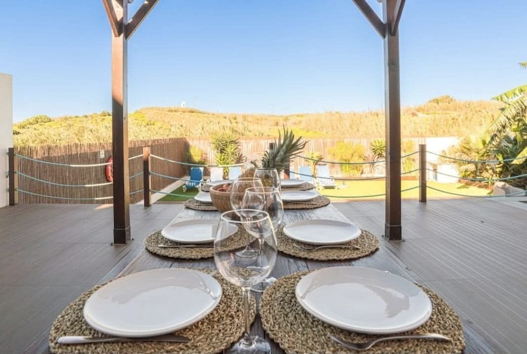 Un espacio de comedor al aire libre con vista a un paisaje natural, con mesas dispuestas con platos blancos y cubertería de vidrio.