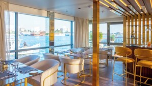 A modern dining area with large windows overlooking a body of water, featuring a bar and seating arrangements. - Sonesta Dahabeya Amirat II - Nile Cruise Ship - 4 nights from Luxor (Luxor)
