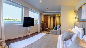 A hotel room with a large bed, a TV, a view of the sea, a small table, and a chair. - Sonesta Dahabeya Amirat II - Nile Cruise Ship - 4 nights from Luxor (Luxor)