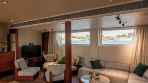 A modern living room with a sofa, chairs, a coffee table, and a TV. Large windows offer a view of the water and cityscape. - Sonesta Dahabeya Amirat II - Nile Cruise Ship - 4 nights from Luxor (Luxor)