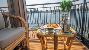 A balcony with a wicker chair, a small table with a teapot, cups, and a plate of food, overlooking a body of water. - Sonesta Dahabeya Amirat II - Nile Cruise Ship - 4 nights from Luxor (Luxor)