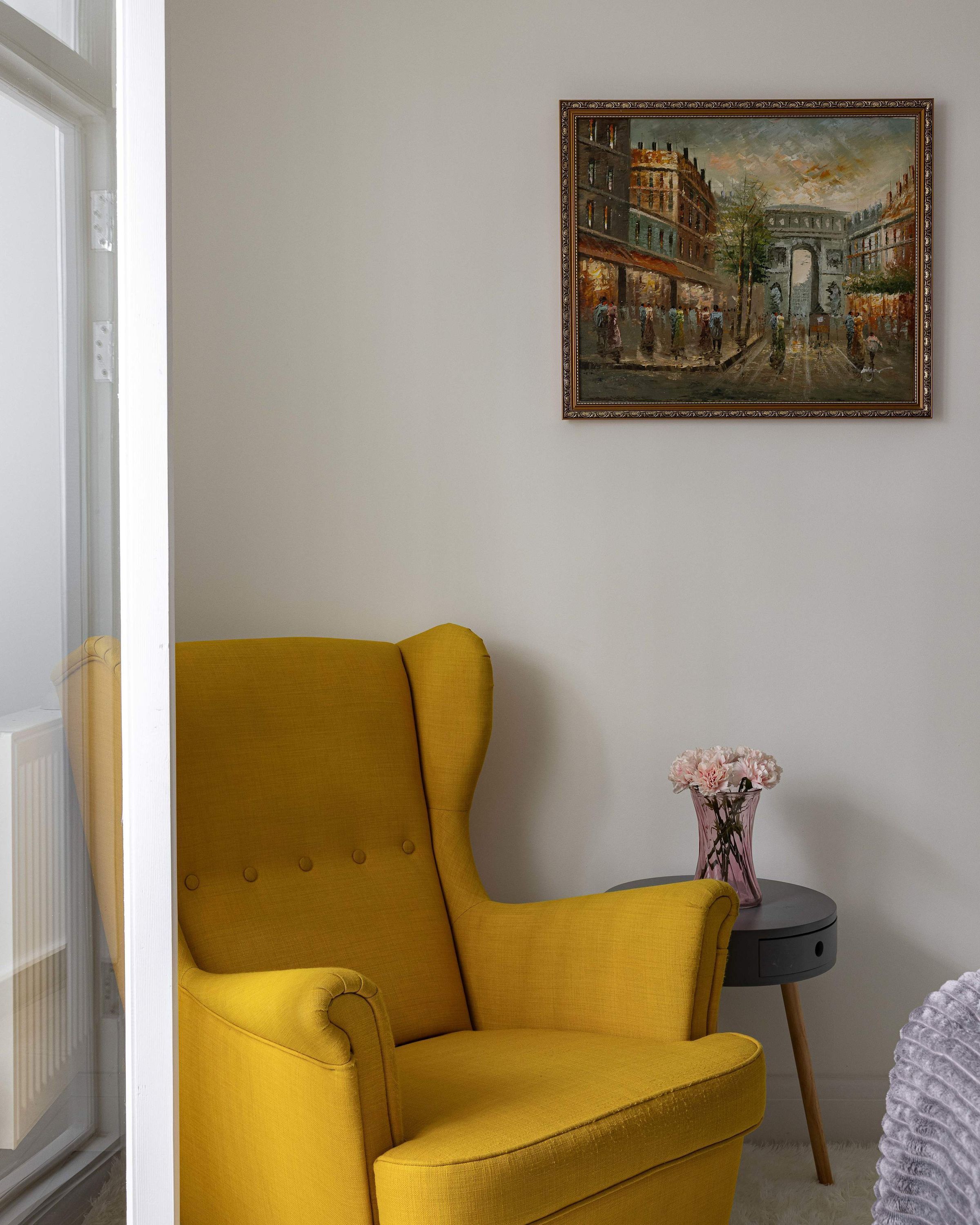 Una stanza con una poltrona color senape, un tavolino rotondo con un vaso di fiori e un quadro appeso al muro.