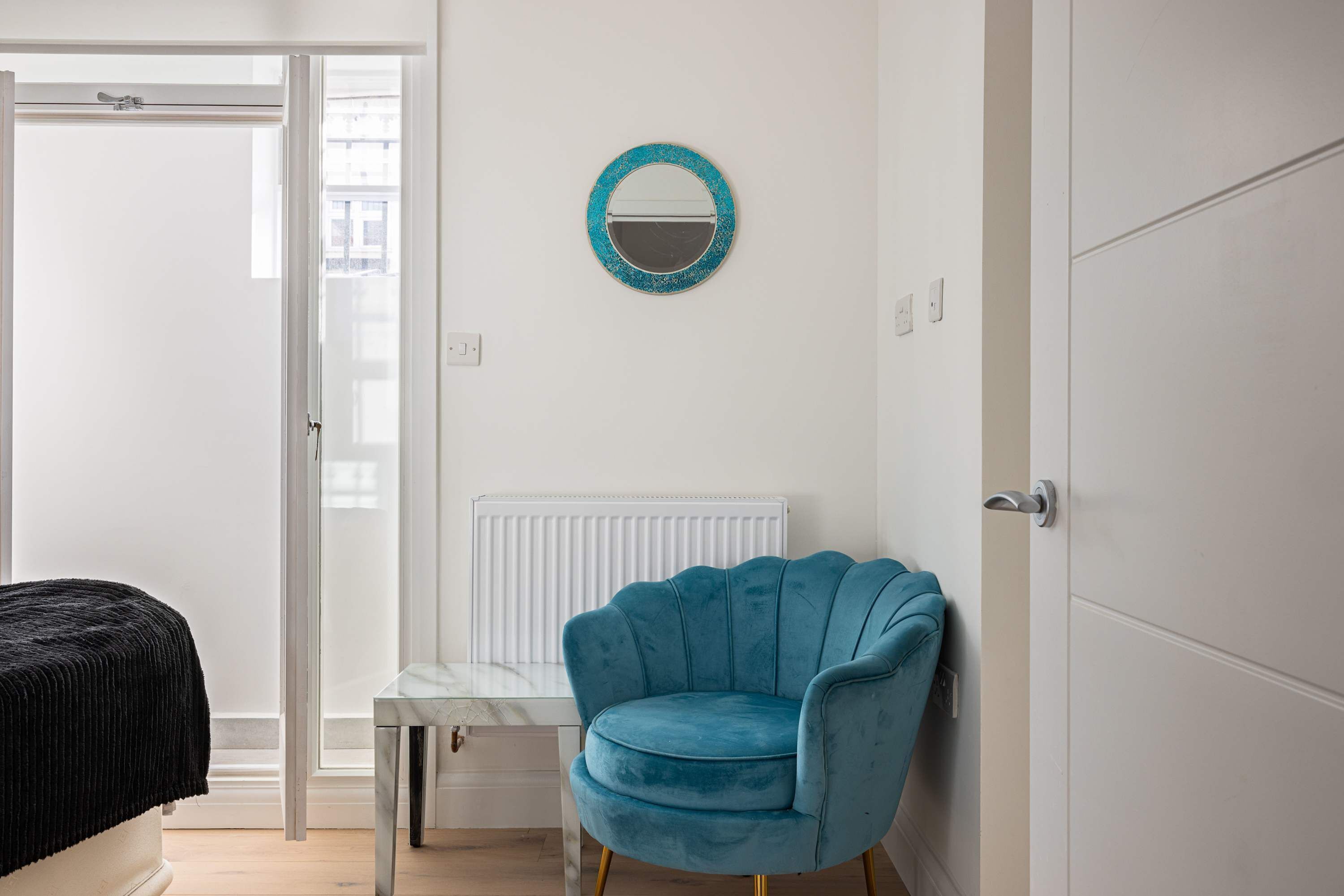 Una stanza con una poltrona blu, un tavolino, un termosifone bianco e una porta che dà su un'altra stanza.