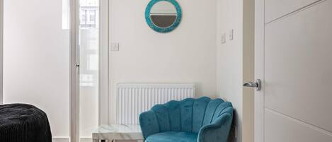 Una stanza con una poltrona blu, un tavolino, un termosifone bianco e una porta che dà su un'altra stanza.