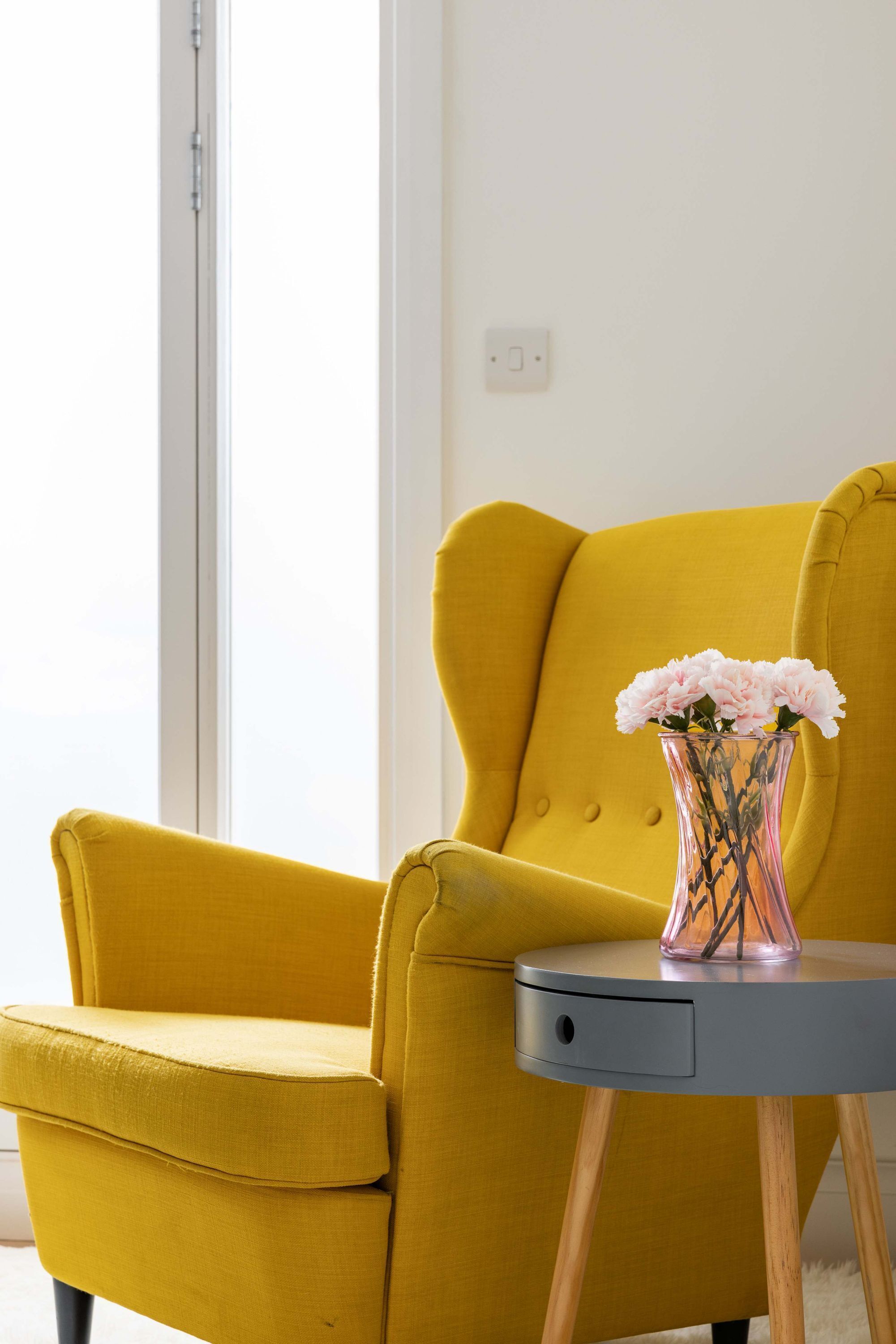 A mustard yellow armchair next to a small round table with a vase of pink flowers.