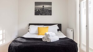 A hotel room with a bed, white and yellow pillows, a black headboard, a black textured bedspread, and a black and white cityscape picture on the wall.
