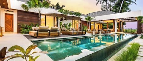 A modern pool area with lounge chairs and umbrellas, surrounded by tropical plants and palm trees.