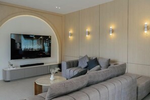 A modern living room with a large flat-screen TV mounted on a curved wall, a sectional sofa, a coffee table, and wall-mounted lighting. - Tierra en Sierra (Pak Chong)