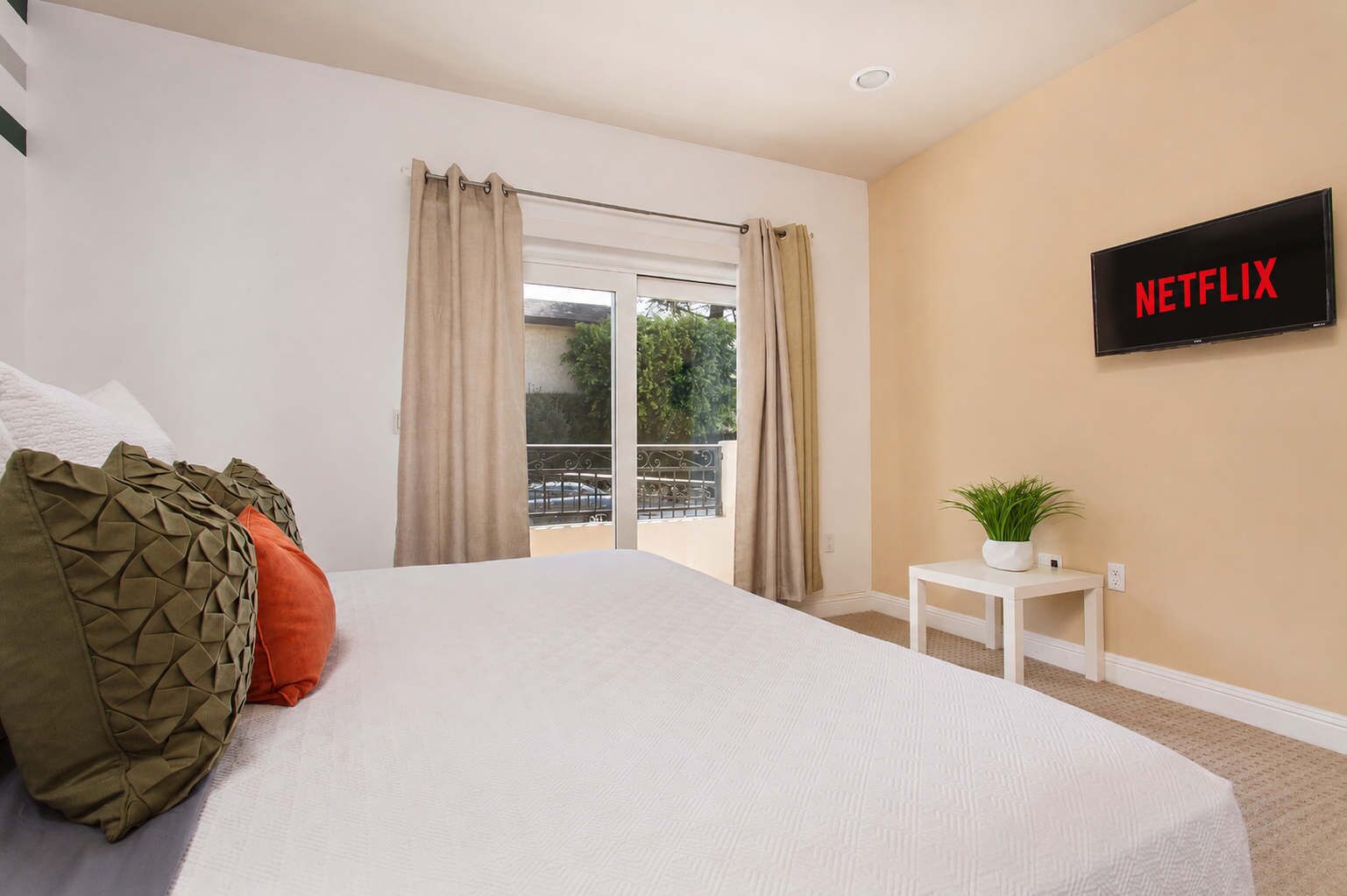 A hotel room with a bed, a TV mounted on the wall, a small table with a plant, and a balcony with curtains.