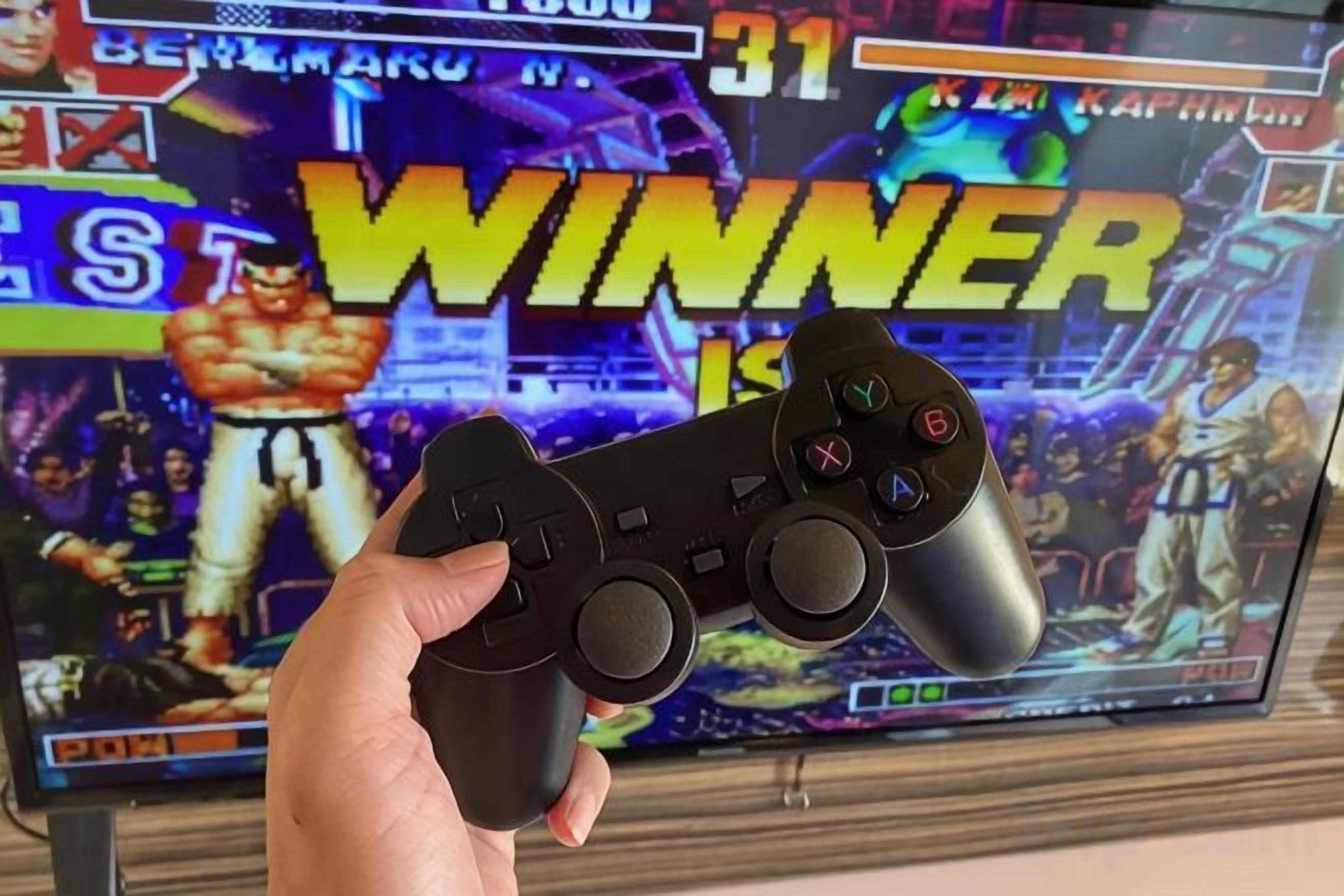 Una mano sostiene un control de videojuegos frente a una pantalla que muestra un juego con la palabra 'WINNER' destacada.