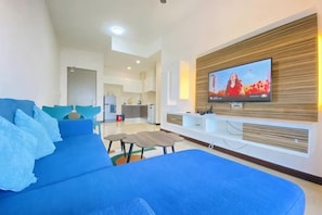 Un salon moderne avec un canapé bleu, un téléviseur à écran plat fixé au mur et une cuisine en arrière-plan.