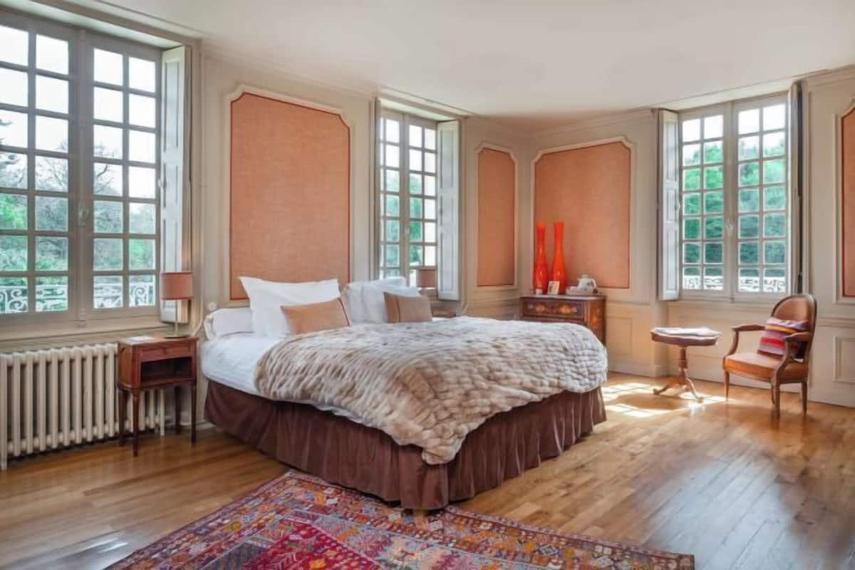 A bedroom with a large bed, a wooden floor, and a window with a view of trees.