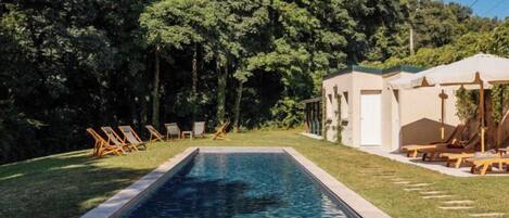 A rectangular swimming pool surrounded by a lawn, with lounge chairs and a building with a door and windows in the background.