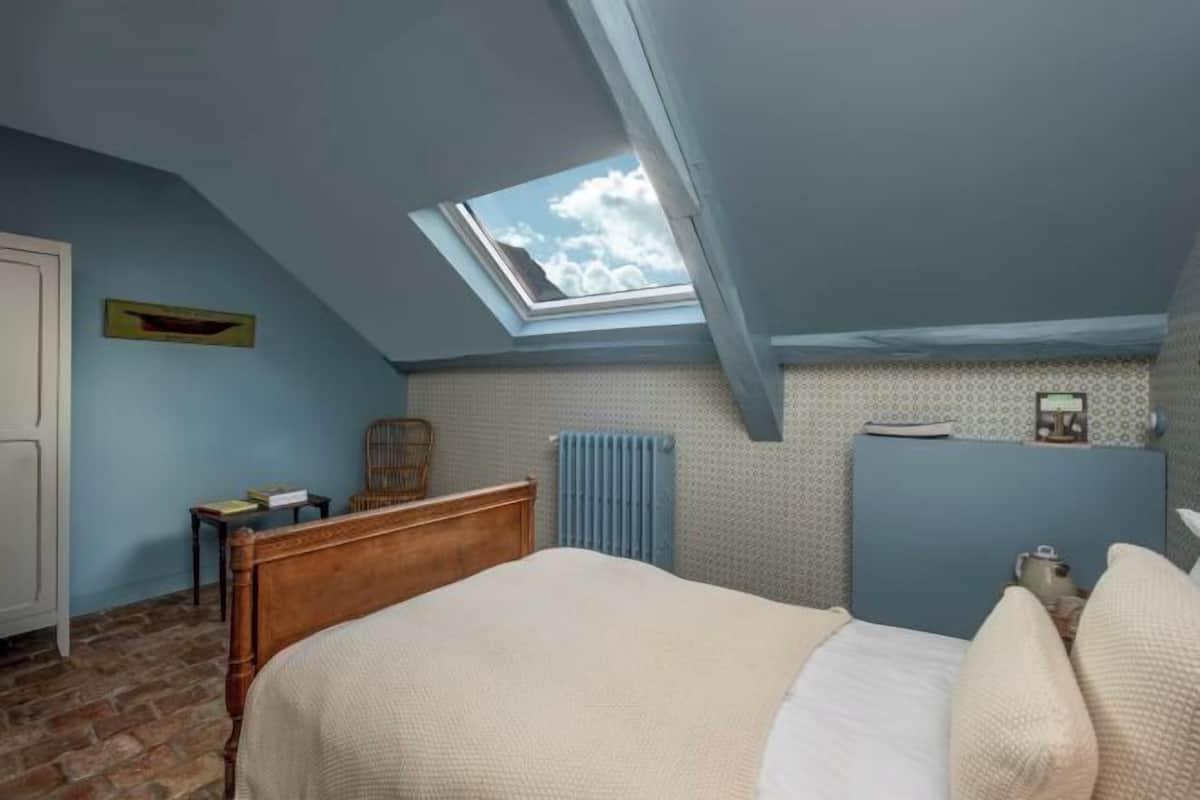 A bedroom with a sloped ceiling, a skylight, a wooden bed, a radiator, and a small table with a chair.