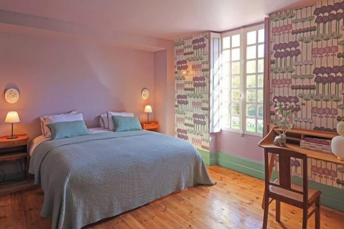 A bedroom with a bed, two bedside tables with lamps, a wooden chair, and a window with patterned curtains.