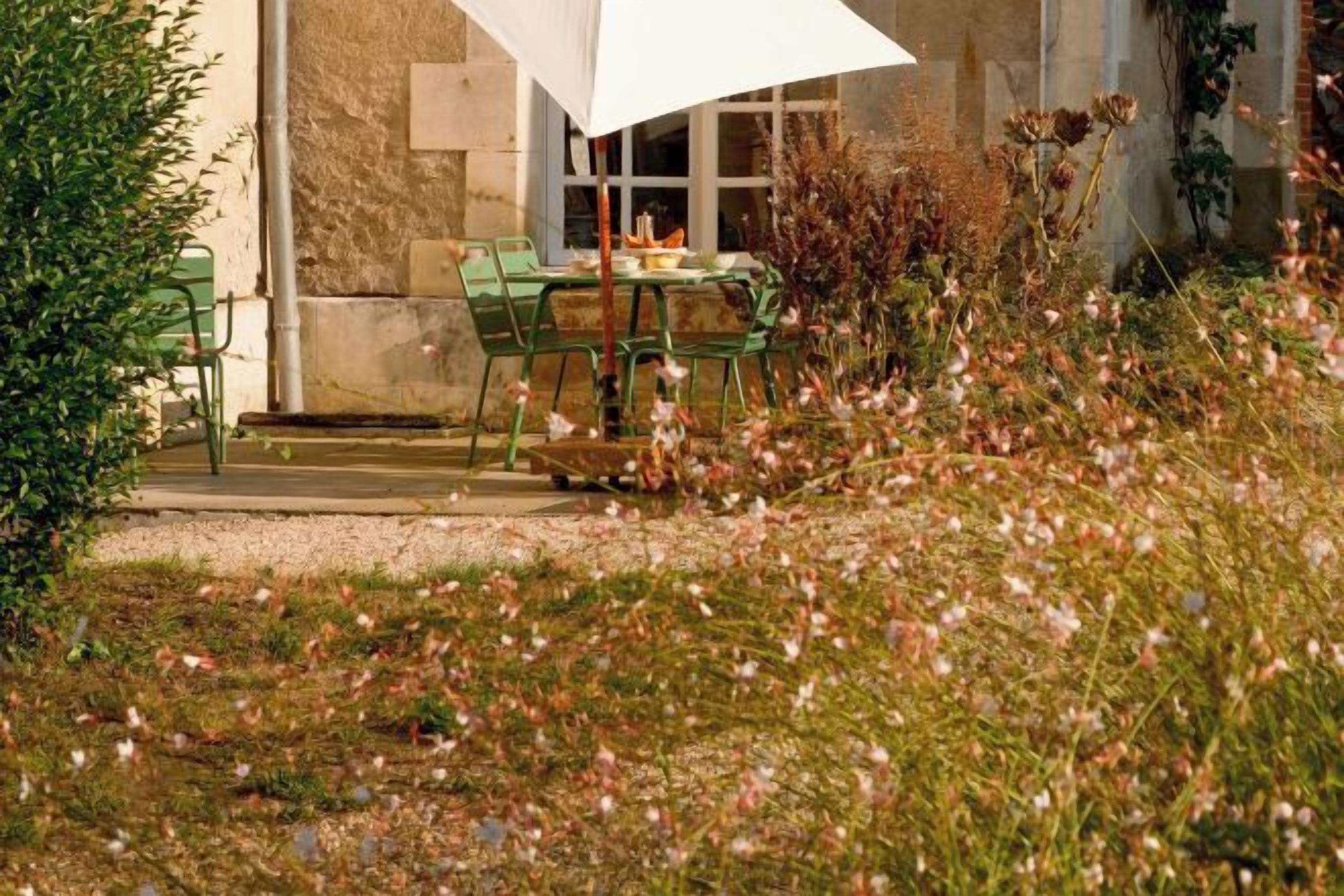 Un área de comedor al aire libre con una mesa y sillas, rodeada por un jardín con flores y un edificio al fondo.