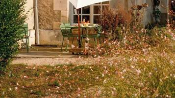 An outdoor dining area with a table and chairs, surrounded by a garden with flowers and a building in the background.