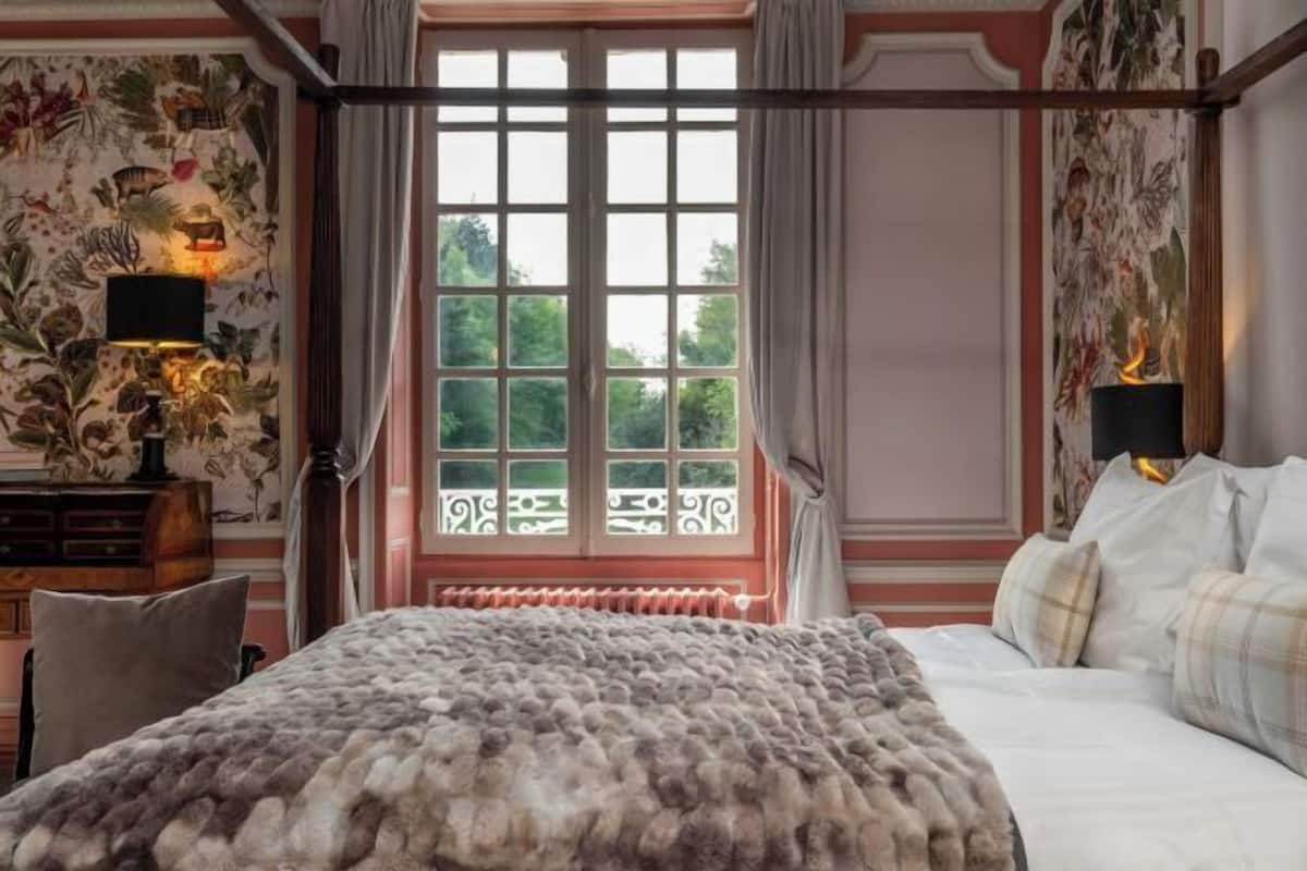 A bedroom with a four-poster bed, floral wallpaper, and a view through a window with a balcony.