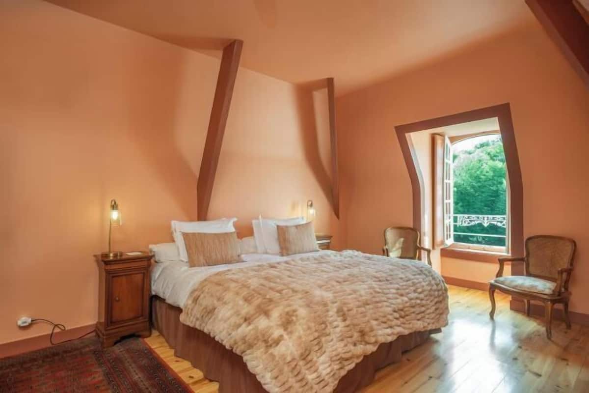A bedroom with a large bed, a nightstand, a chair, and a window with a view of greenery.