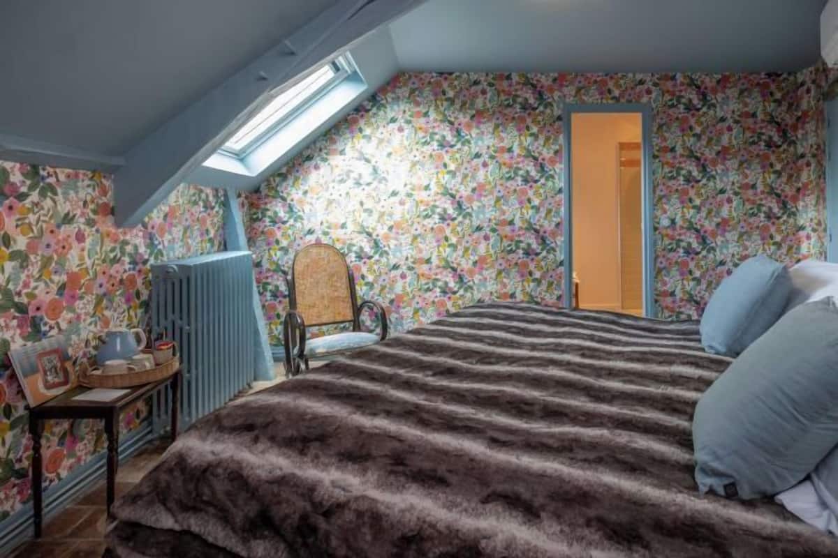 A bedroom with a skylight, floral wallpaper, a bed with a striped blanket, a chair, and a radiator.