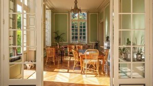A dining area with a wooden floor, wicker chairs, a chandelier, and a large window with multiple panes. - Huberdière Castle (Nazelles-Negron)