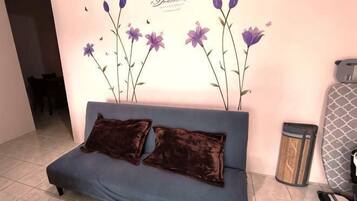 A blue sofa with two dark cushions, a wall with purple flowers, and a trash can.