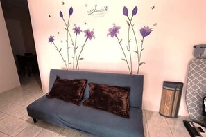 A blue sofa with two dark cushions, a wall with purple flowers, and a trash can. - TAWAU COZY HOMESTAY @ MEGAH JAYA 10 (Tawau)