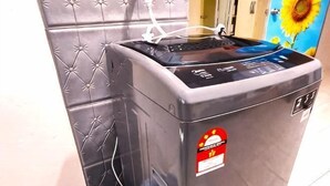 A gray washing machine with a control panel, a red sticker, and a blue bag on the floor.