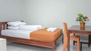 A single bed with white linens, a brown blanket, and a rolled towel on the side. There is a wooden table with a woven basket containing a plant. - Amor Homestay near Eltari International Airport Mitra RedDoorz (Liliba)