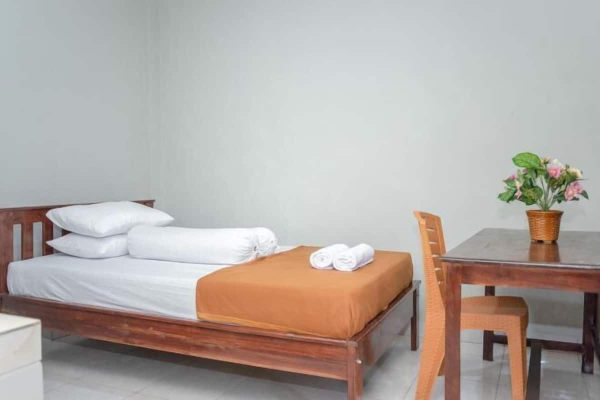 Una cama individual con sábanas blancas, una manta marrón y una toalla enrollada al lado. Hay una mesa de madera con una cesta tejida que contiene una planta.