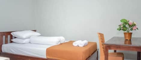 Una cama individual con sábanas blancas, una manta marrón y una toalla enrollada al lado. Hay una mesa de madera con una cesta tejida que contiene una planta.