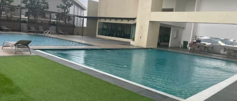 Piscina con césped y sillas de descanso, rodeada por un edificio con amplias ventanas y un balcón.