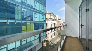 Un edificio moderno con amplios ventanales, un balcón con barandilla de vidrio y vistas a edificios tradicionales abajo.