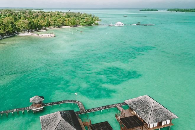 An aerial view of a tropical resort with overwater bungalows connected by walkways, clear turquoise water, and lush greenery.