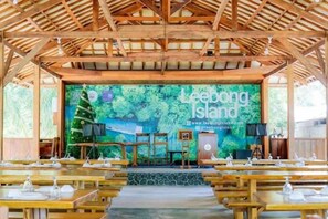 A covered outdoor dining area with a stage in the background, tables set with white tableware, and a banner reading 'Leebong Island.' - Leebong Island (Pegantungan)