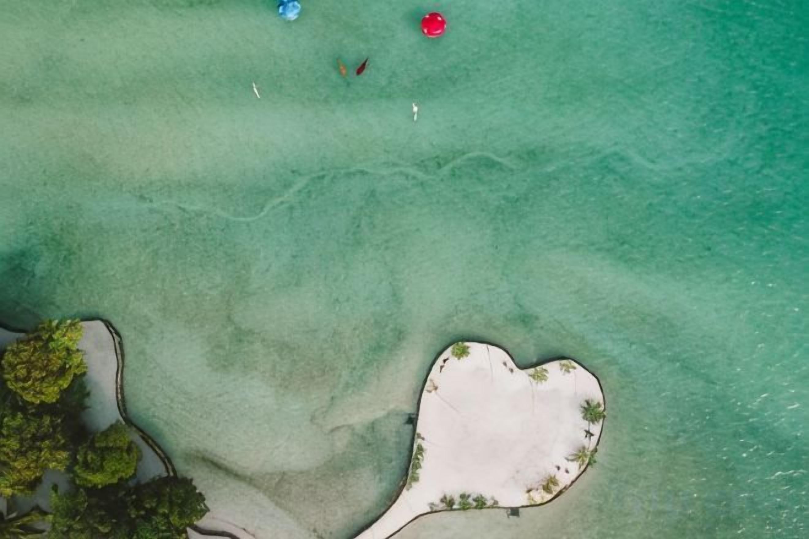 An aerial view of a beach with clear turquoise water, a heart-shaped sandbar, and sparse vegetation.