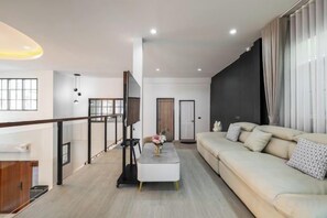 A modern living room with a sofa, a coffee table, and a TV. There is a staircase with a railing and a door in the background. - Luxury Private Pool Villa "Psalms 23 Residence @Huaikwang" (Bangkok)