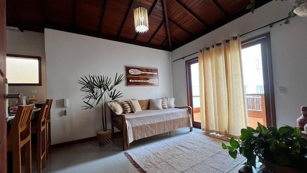 A cozy living room with a sofa, a dining table, and chairs. There is a large window with curtains, a ceiling fan, and a plant. - Apartamento Novo ao lado da Rua Mucugê (Porto Seguro)