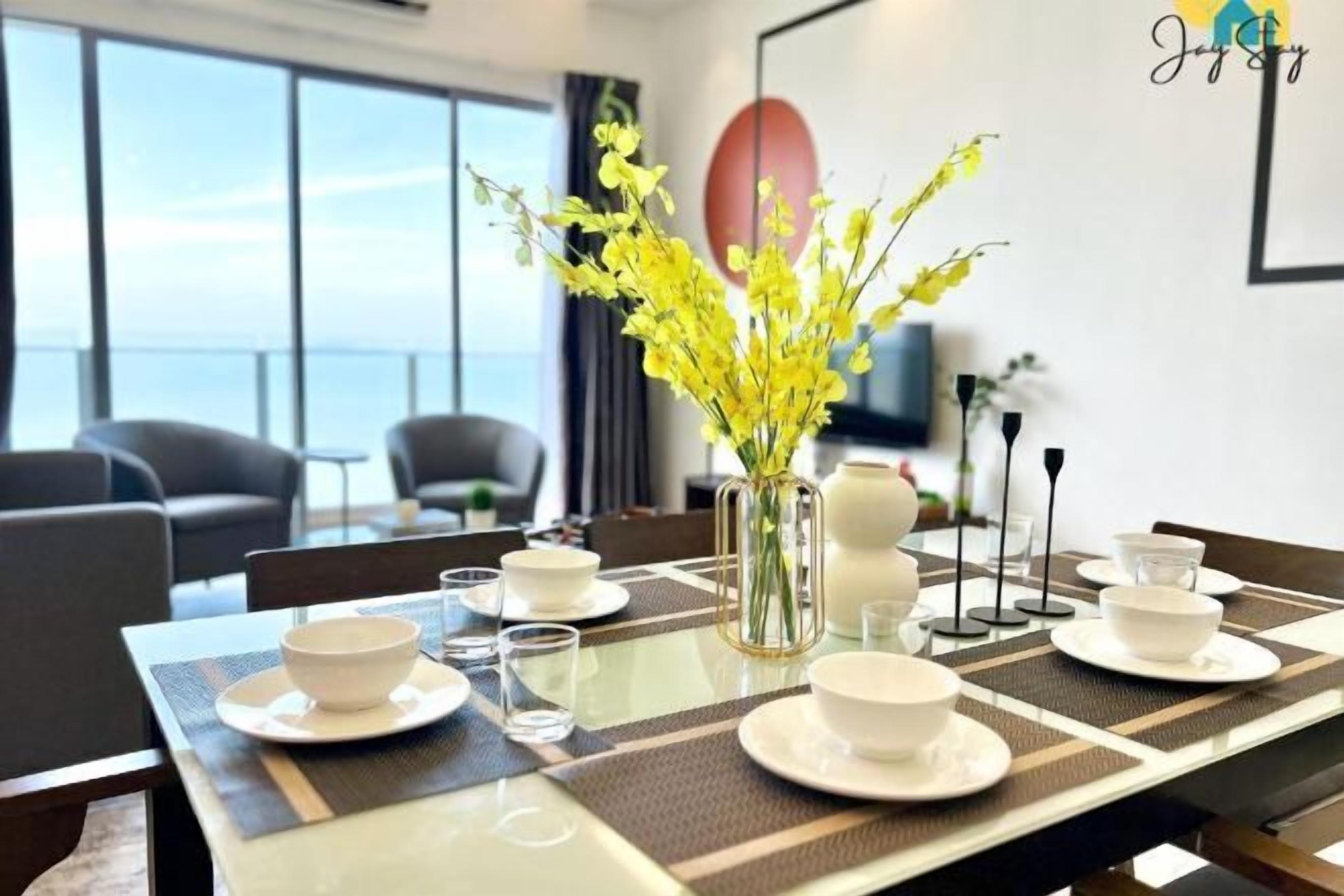 Un coin repas avec une table dressée pour quatre personnes, un vase rempli de fleurs jaunes et une vue sur la mer, visible à travers de grandes fenêtres.