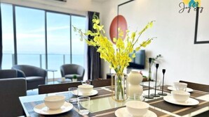 A dining area with a table set for four, a vase with yellow flowers, and a view of the sea through large windows. - SilverScape Melaka/ 3BR/ 6-10 pax/Bathtub/Seaview/ Infinity Pool/ Jonker St (Melaka)