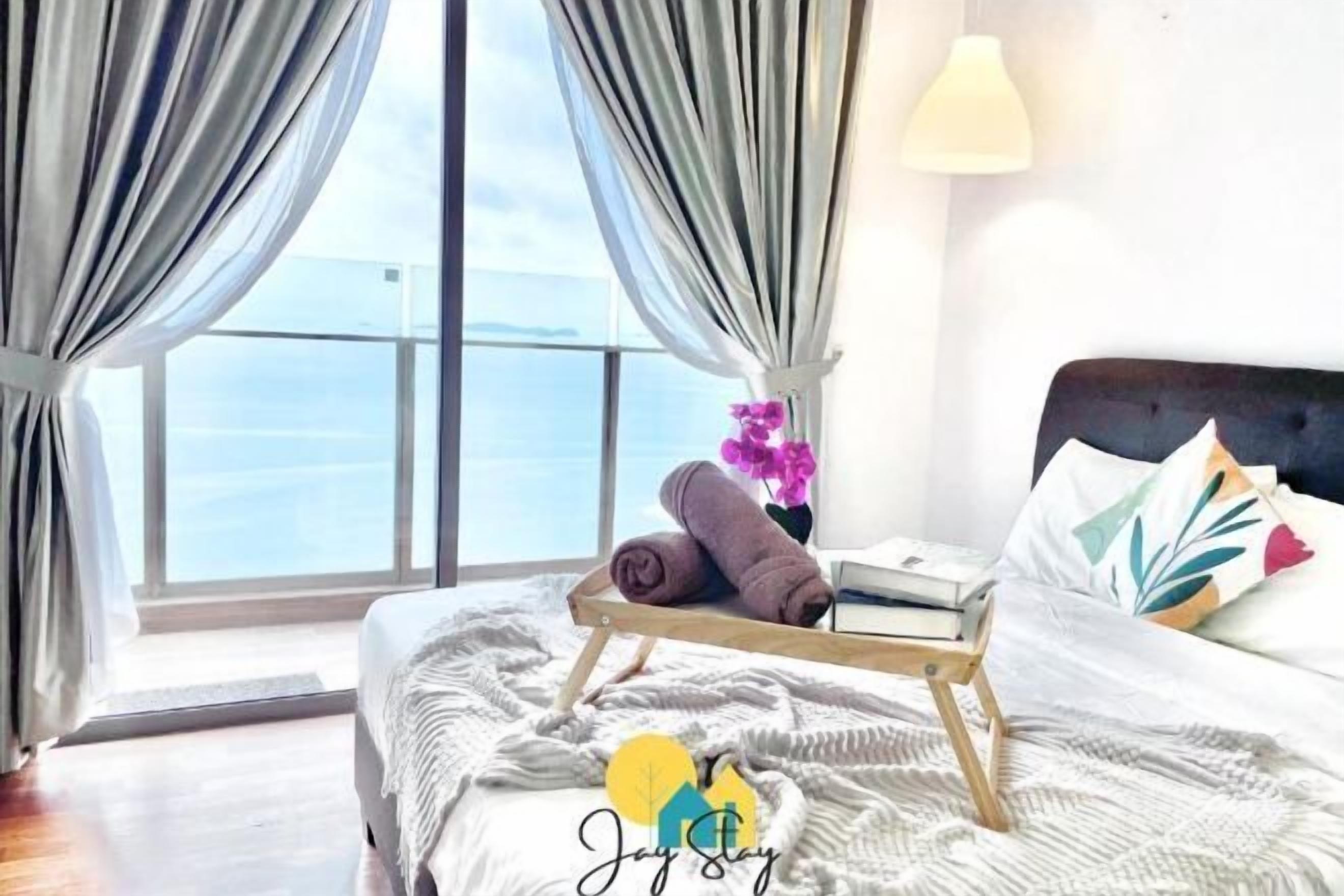 Une chambre à coucher avec un lit, une table de chevet sur laquelle se trouvent des serviettes et des livres, une fenêtre avec des rideaux et une vue sur la mer.