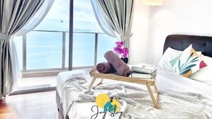 A bedroom with a bed, a bedside table with towels and books, a window with curtains, and a view of the sea. - SilverScape Melaka/ 3BR/ 6-10 pax/Bathtub/Seaview/ Infinity Pool/ Jonker St (Melaka)