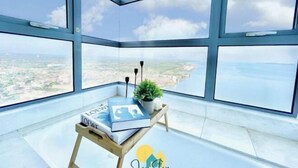 A high-rise room with large windows, a small wooden table with a book and a plant, and a clear view of the cityscape and coastline. - SilverScape Melaka/ 3BR/ 6-10 pax/Bathtub/Seaview/ Infinity Pool/ Jonker St (Melaka)