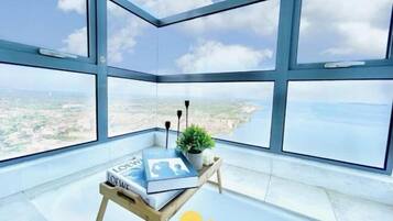 Une chambre située dans un immeuble de grande hauteur, dotée de grandes fenêtres, d’une petite table en bois sur laquelle se trouvent un livre et une plante, et offrant une vue dégagée sur le paysage urbain et la côte.